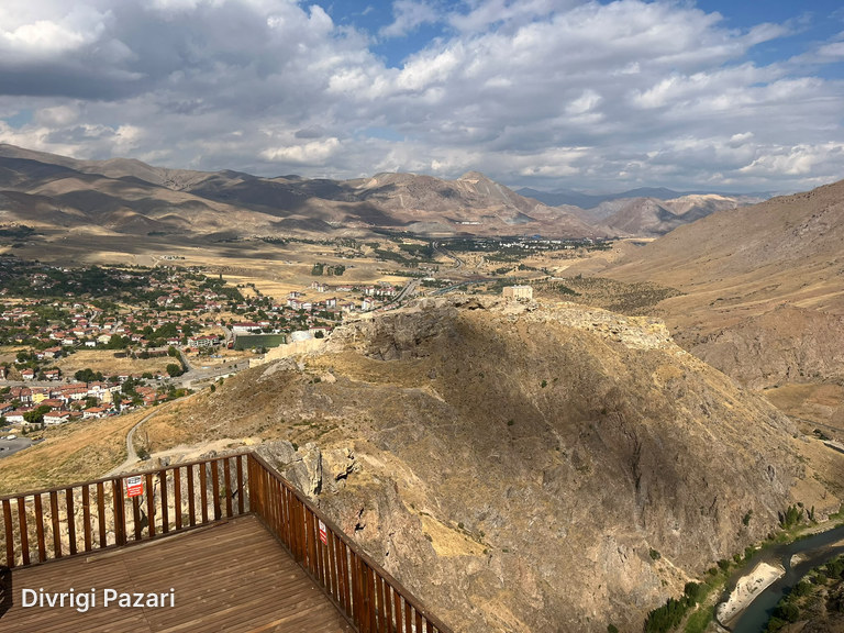 Beyond a wooden viewing deck: a rocky ridge, the houses of Divriği, the valley and a cloudy sky