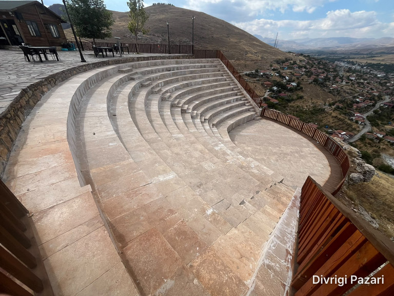 Curved stone seating steps, a wooden building and tables; beyond the railing on the right, the houses of Divriği