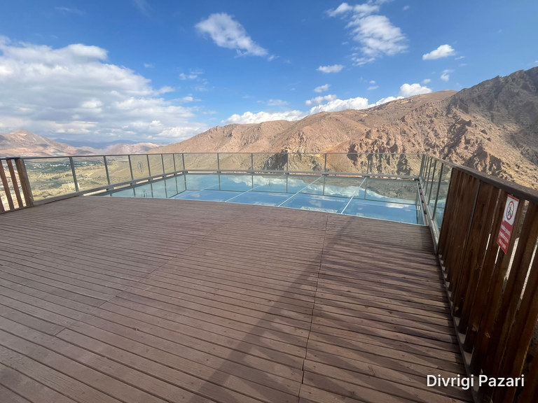 A glass-floored platform with a glass balustrade at the end of the wooden deck; rocky mountains behind