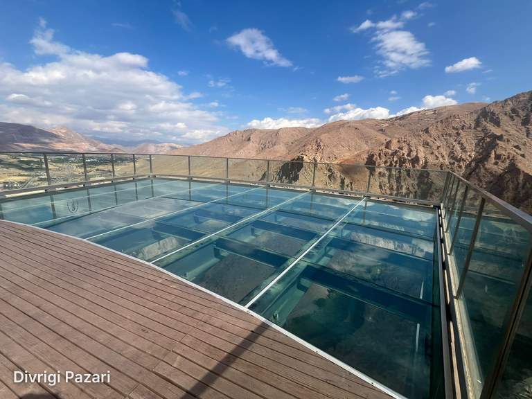 Clouds reflected in the glass floor of the viewing terrace, with the wooden deck in front and rocky mountains behind