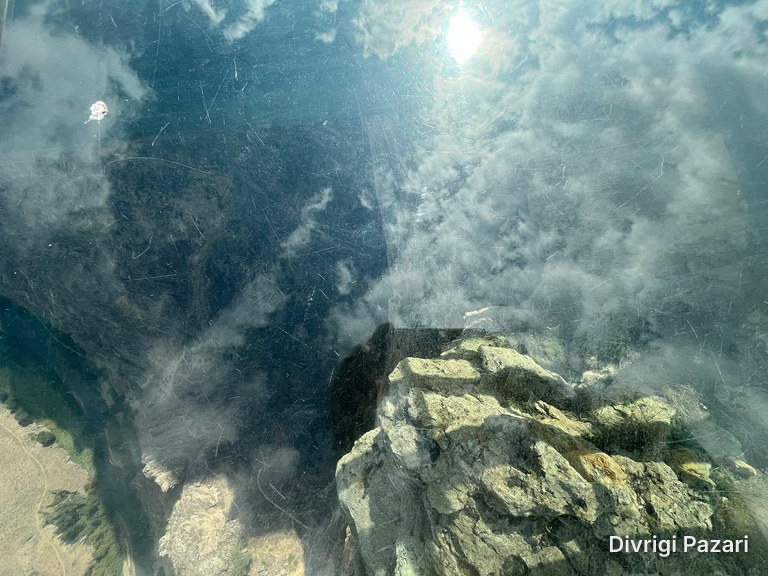 Looking down through the glass floor: clouds and the sun reflected in the glass, rocks below