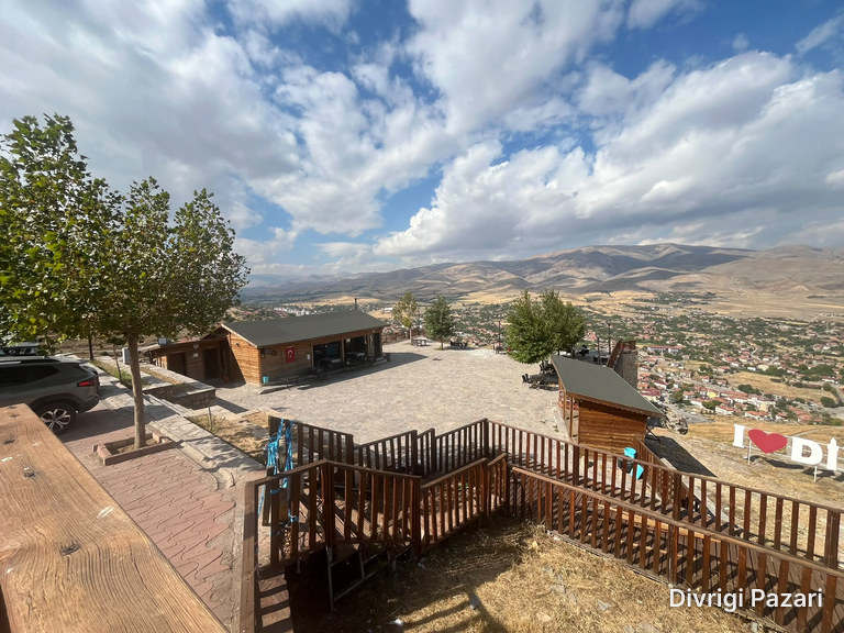A stone-paved square, wooden café buildings and trees; the houses of Divriği and the mountains beyond
