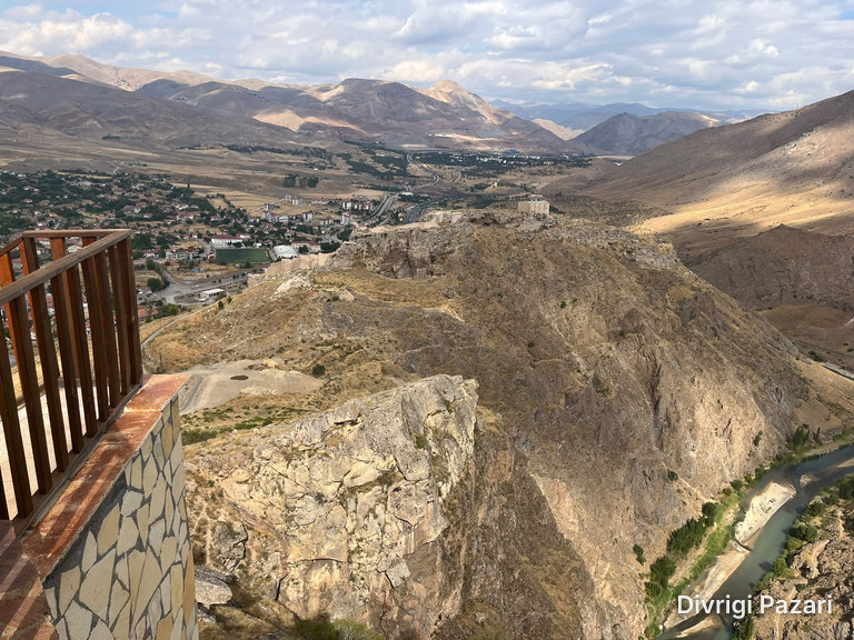 Beyond a wooden railing on a stone wall: a rocky ridge, the houses of Divriği and the Çaltı below