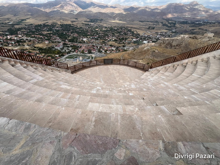 Curved stone seating steps; beyond the railing, the roofs and gardens of Divriği and the mountains