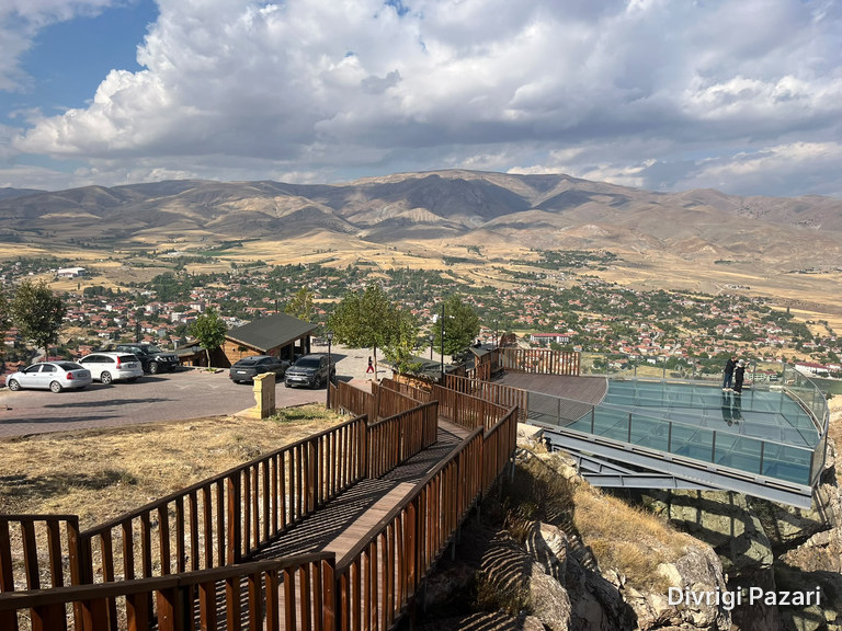A wooden walkway down the rocks and the glass terrace reaching out over the drop; a car park on the left, the town and mountains behind