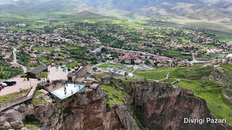 From above: the glass terrace at the cliff edge, the square and the houses of Divriği; green slopes