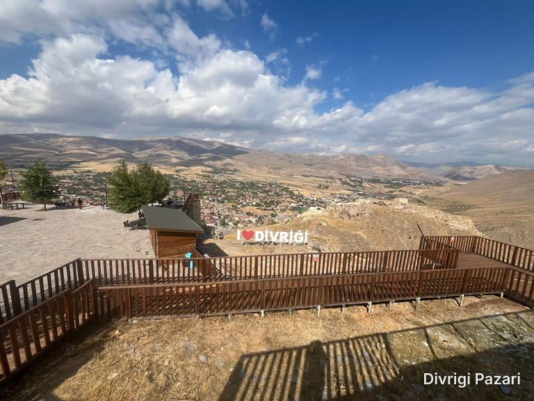 Walkways with wooden railings, the Divriği sign and a wooden hut; the town and mountains beyond