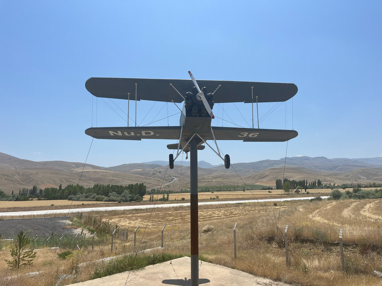 The monument model of the Nu.D.36 aircraft raised on a pylon at the campus, with Divriği's fields and mountains behind