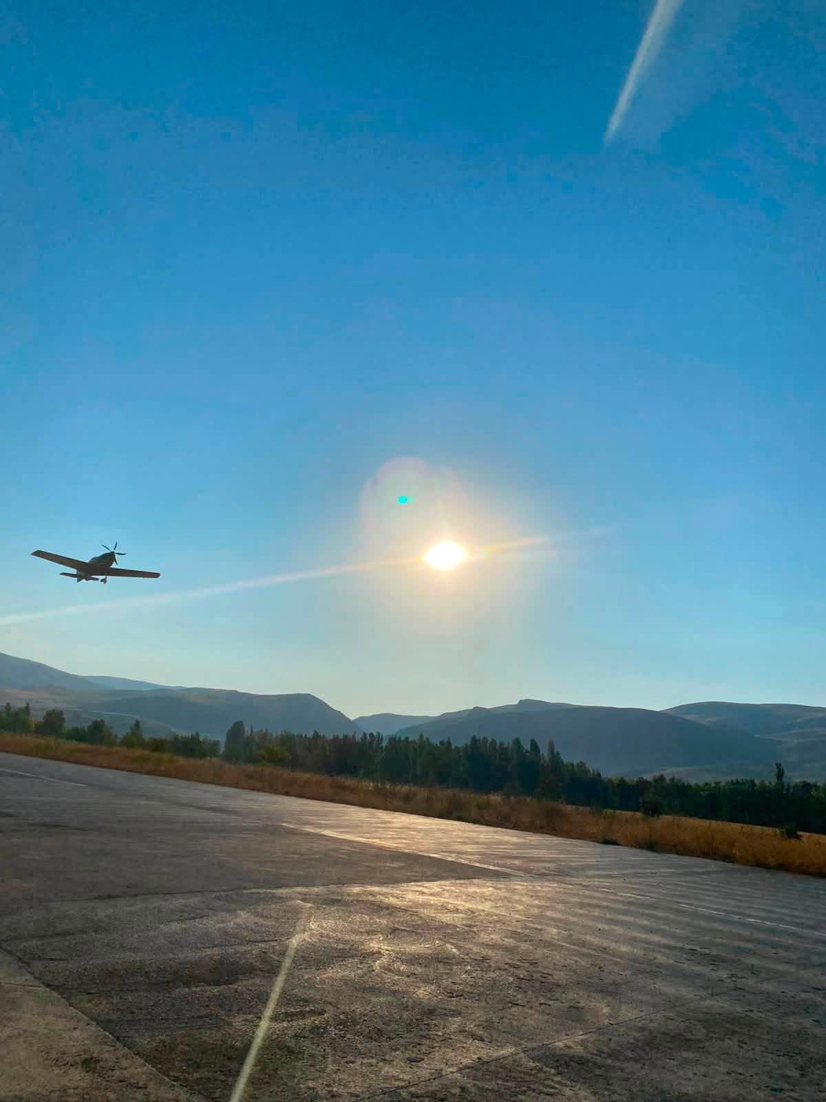 An aircraft in flight over the runway at Nuri Demirağ Air Campus, the concrete strip in the foreground and the sun and mountains behind